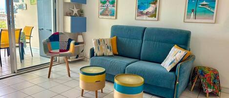 Una sala de estar con un sofá azul, dos mesitas redondas, una silla y un ventanal de vidrio que da a un balcón.