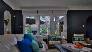 Sala de estar com sofá branco, almofadas azuis e verdes, mesa de centro com laranjas e vista pela janela com treliça.