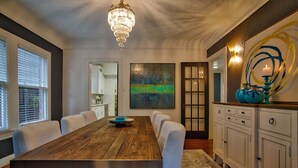 Uma sala de jantar com mesa de madeira, cadeiras brancas, um lustre e um quadro na parede.