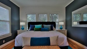 Quarto com uma cama grande, duas mesas de cabeceira, um banco e três janelas com persianas.