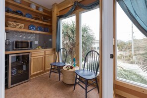 A kitchen with wooden cabinets, a microwave, and a wine cooler. There are two chairs and a small table with a bottle and glasses.