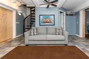 A living room with a grey sofa, a brown rug, a ceiling fan, and a painting of flowers on the wall. - Tower Four - Oceanfront Historic Missile Tower - 3 BR / 2.5 BATH (Surf City)