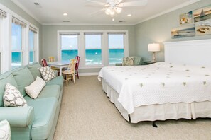 A bedroom with a bed, a sofa, a table, chairs, and a view of the ocean.