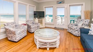 A living room with a glass-top coffee table, white wicker furniture, a TV, and a view of the ocean through large windows. - Vitamin Sea - Oceanfront, 5 BR Home in Surf City - Elevator! (Surf City)