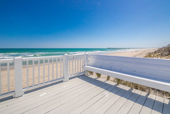 A wooden deck with a white railing overlooking a beach and a pier in the distance.