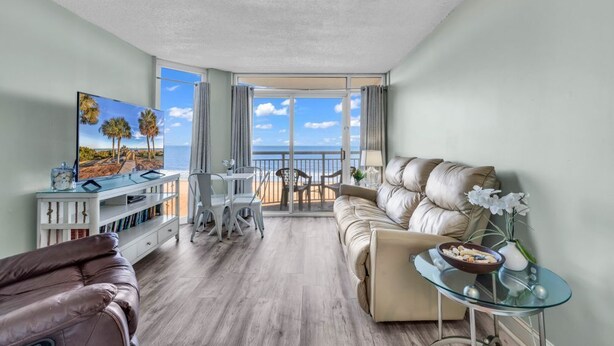Ein modernes Wohnzimmer mit einer Ledersofa, einem Glastisch und einem Fernseher auf einer weißen Ablage. Durch die Glastüren ist ein Balkon mit Meerblick zu sehen.