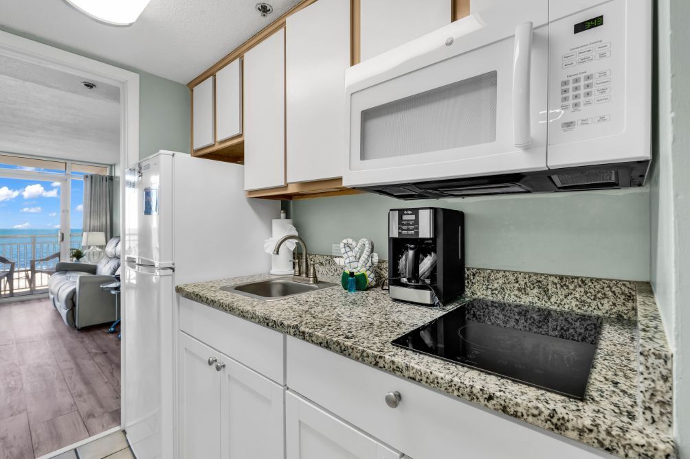 A kitchen with white cabinets, a granite countertop, a microwave, a coffee maker, and a view of the ocean.