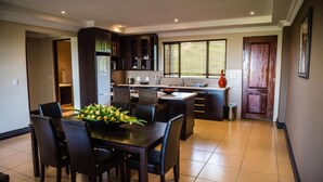 A modern kitchen with dark cabinetry, a dining area with a table and chairs, and a large window with blinds. - The Royal Solwezi Hotel (Solwezi)