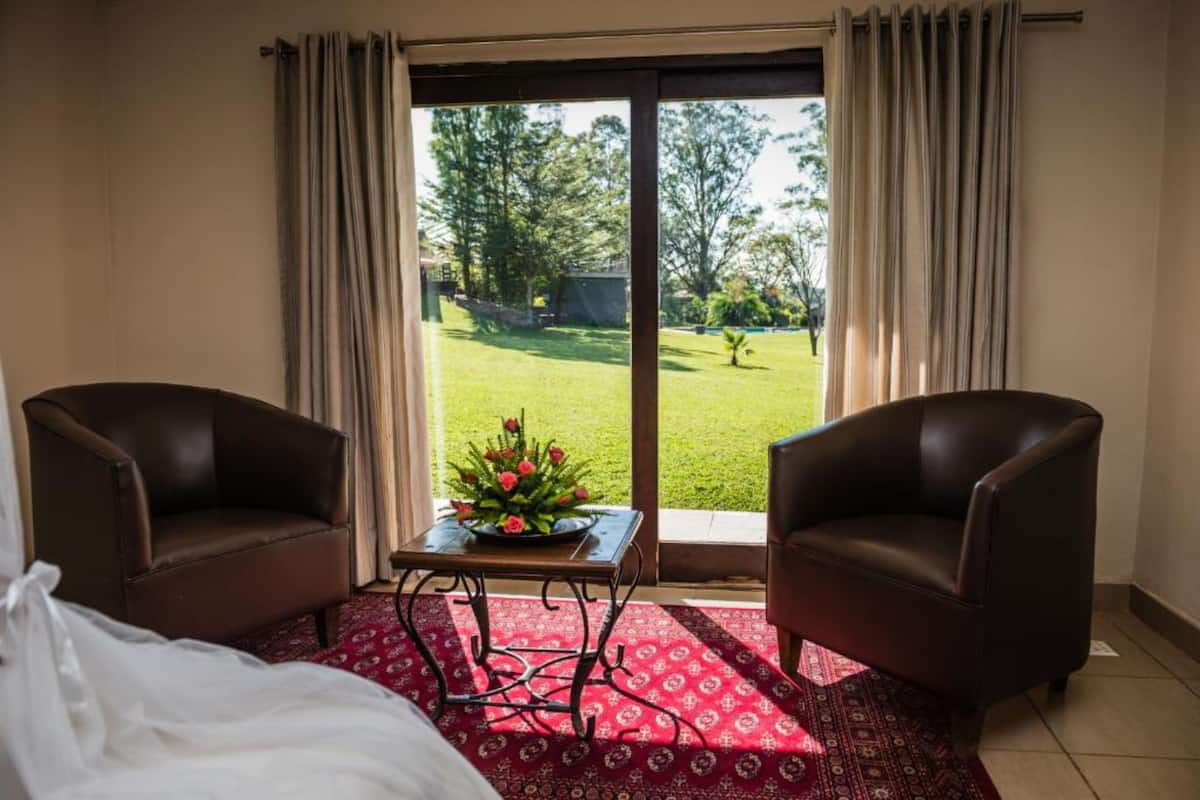 A hotel room with a bed, two leather armchairs, a small table with a flower arrangement, and a view of a green lawn through a sliding glass door.