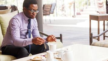 Une personne est assise à une table, devant une théière, des tasses et une assiette de nourriture, dans un intérieur bien éclairé.