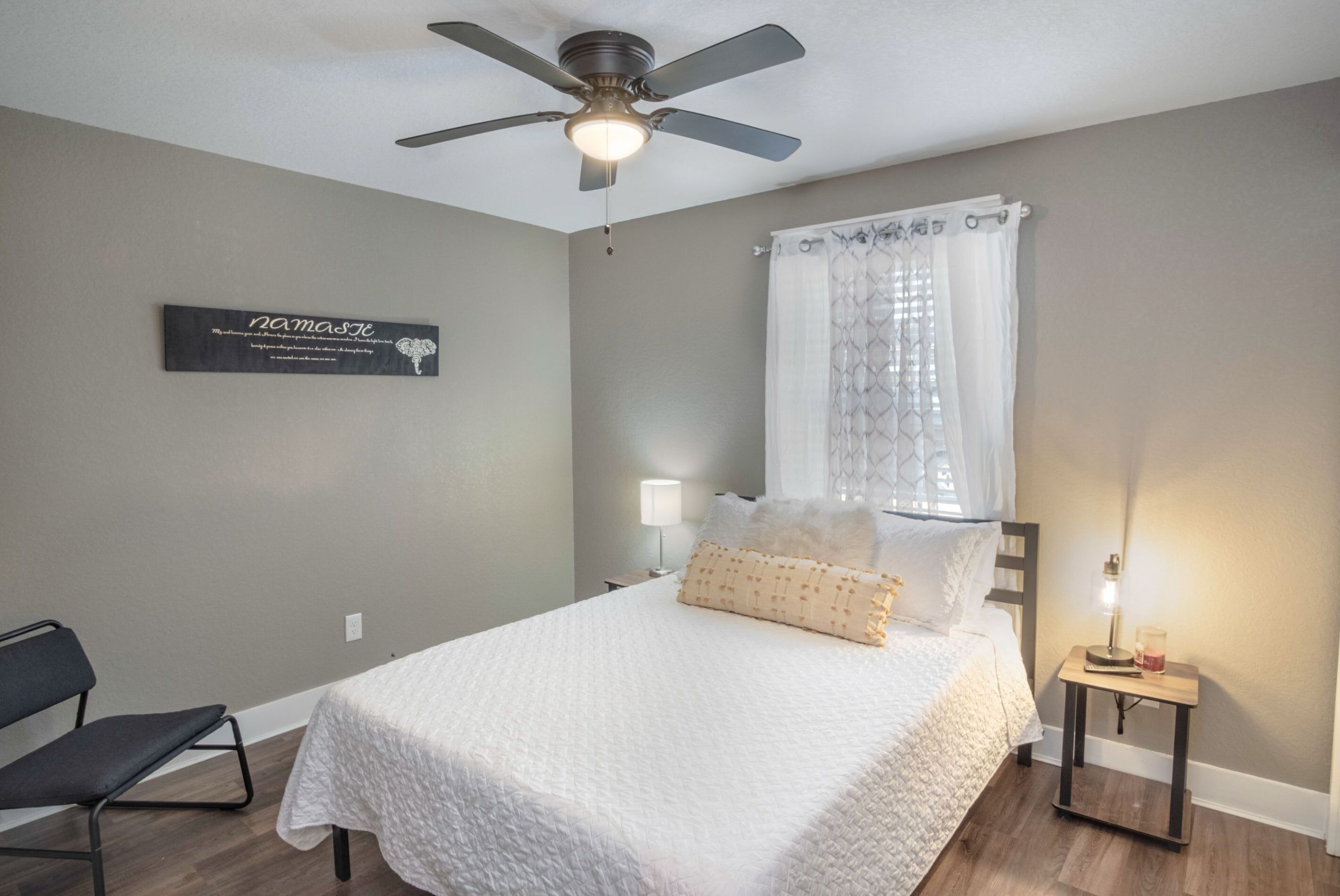 A bedroom with a bed, a ceiling fan, a nightstand with a lamp, a chair, and a wall-mounted sign.