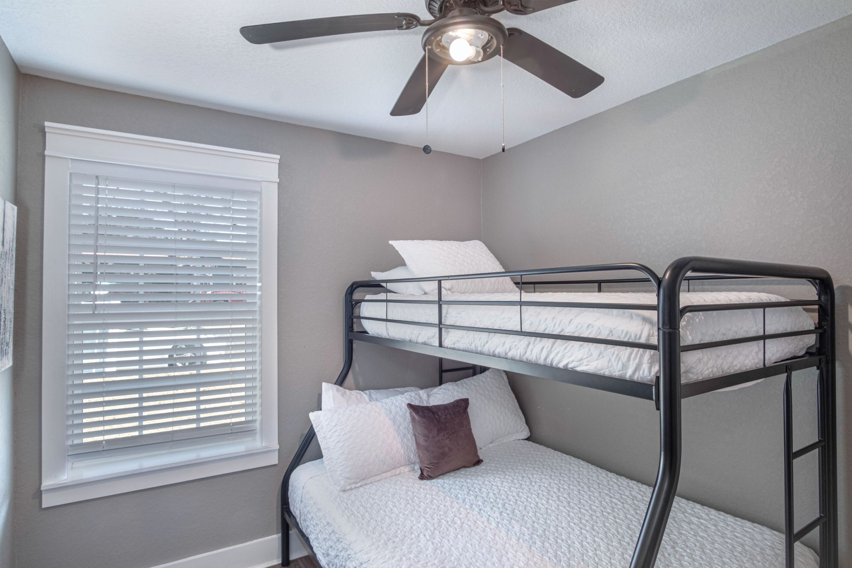 A bunk bed room with a ceiling fan, a window with blinds, and a wall-mounted picture.