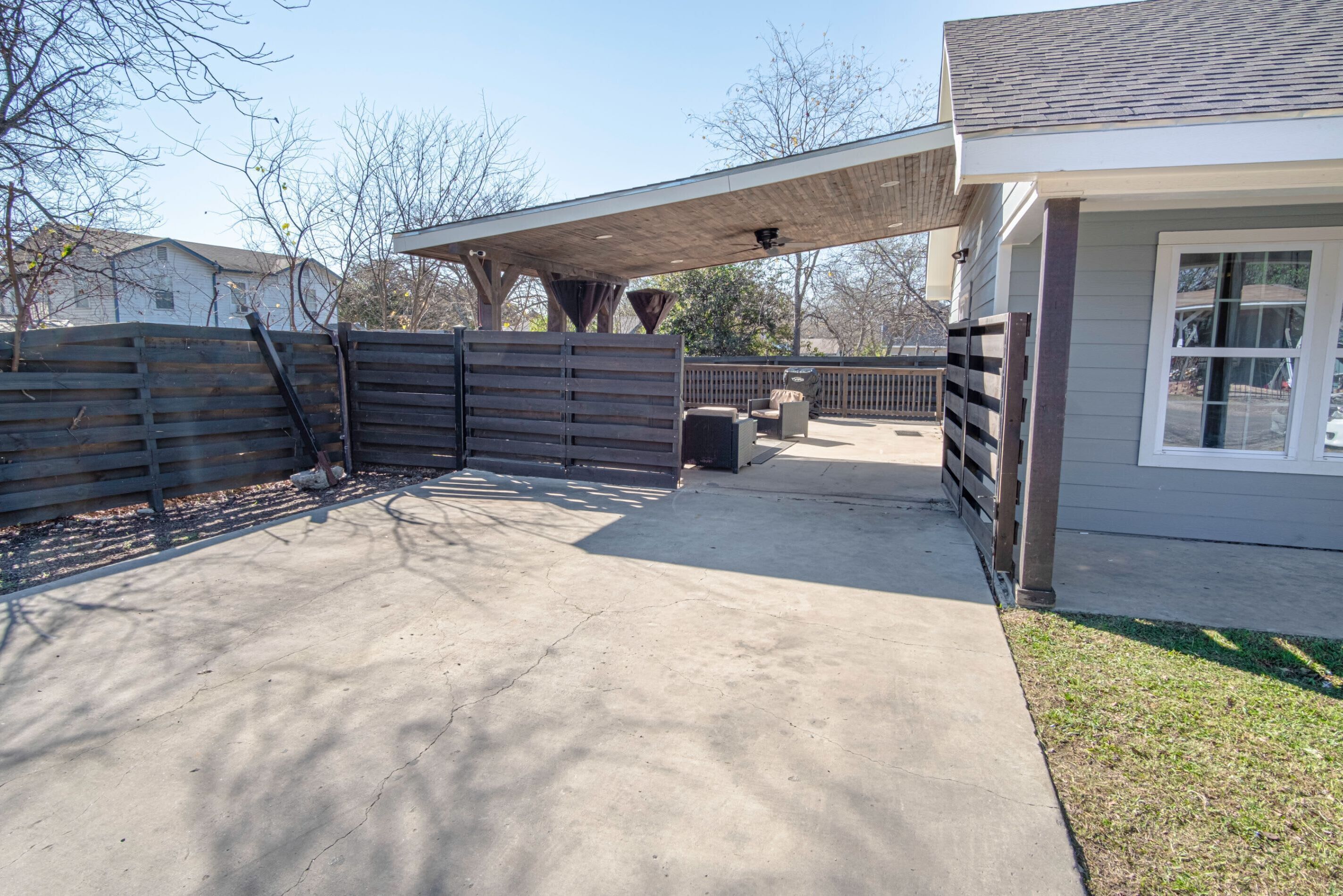 A covered patio area with a concrete floor, a wooden fence, and a building with a window.