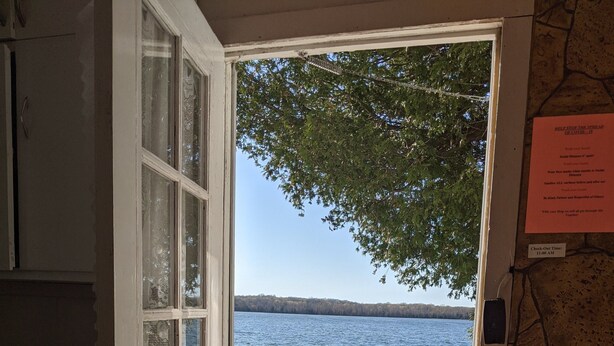 A wooden deck leading to a lake with a picnic table, a stone wall, and a door with a check-out sign. - Lakeview Scenic Cabin sleep 4 (Mar)