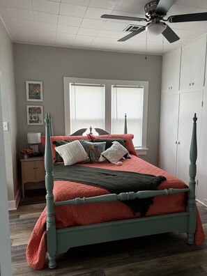 A bedroom with a four-poster bed, a ceiling fan, a window with blinds, and a nightstand with a lamp.