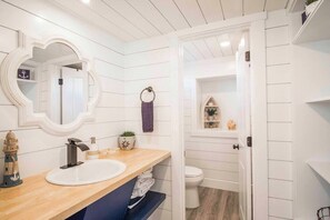 Un baño con tocador de madera, un espejo redondo, un lavamanos blanco y un inodoro.