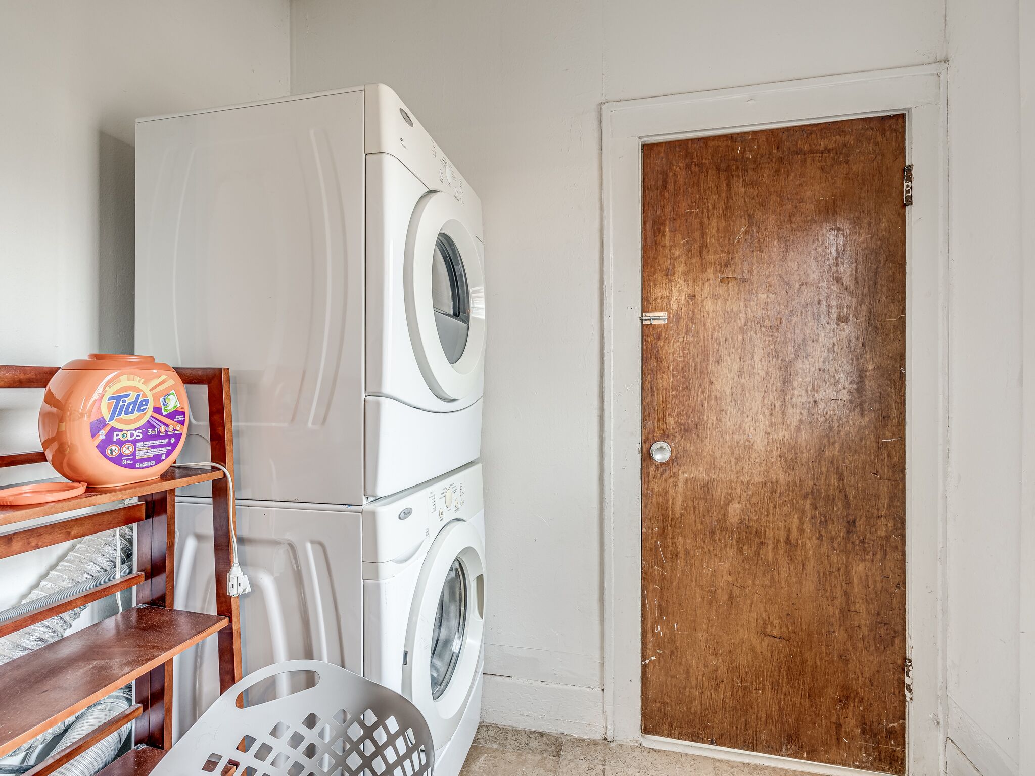 A laundry area with a stacked washer and dryer, a Tide PODS container, and a wooden door.