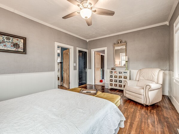 A bedroom with a large bed, a chair, a dresser with pictures, a ceiling fan, and a door leading to another room. - The Frida House in the Urban Core (Oklahoma City)