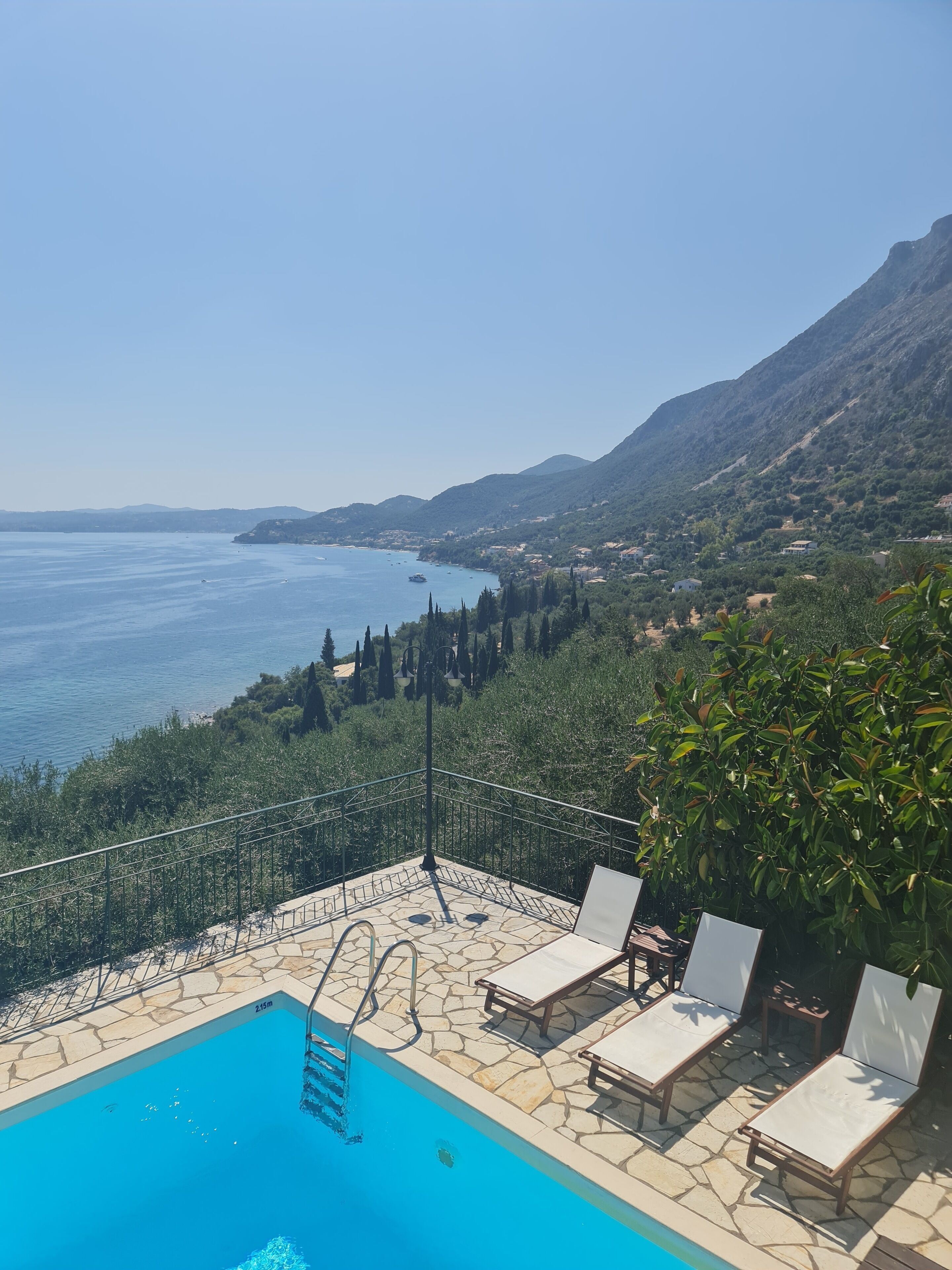A swimming pool with lounge chairs, a stone deck, and a view of the sea and mountains.