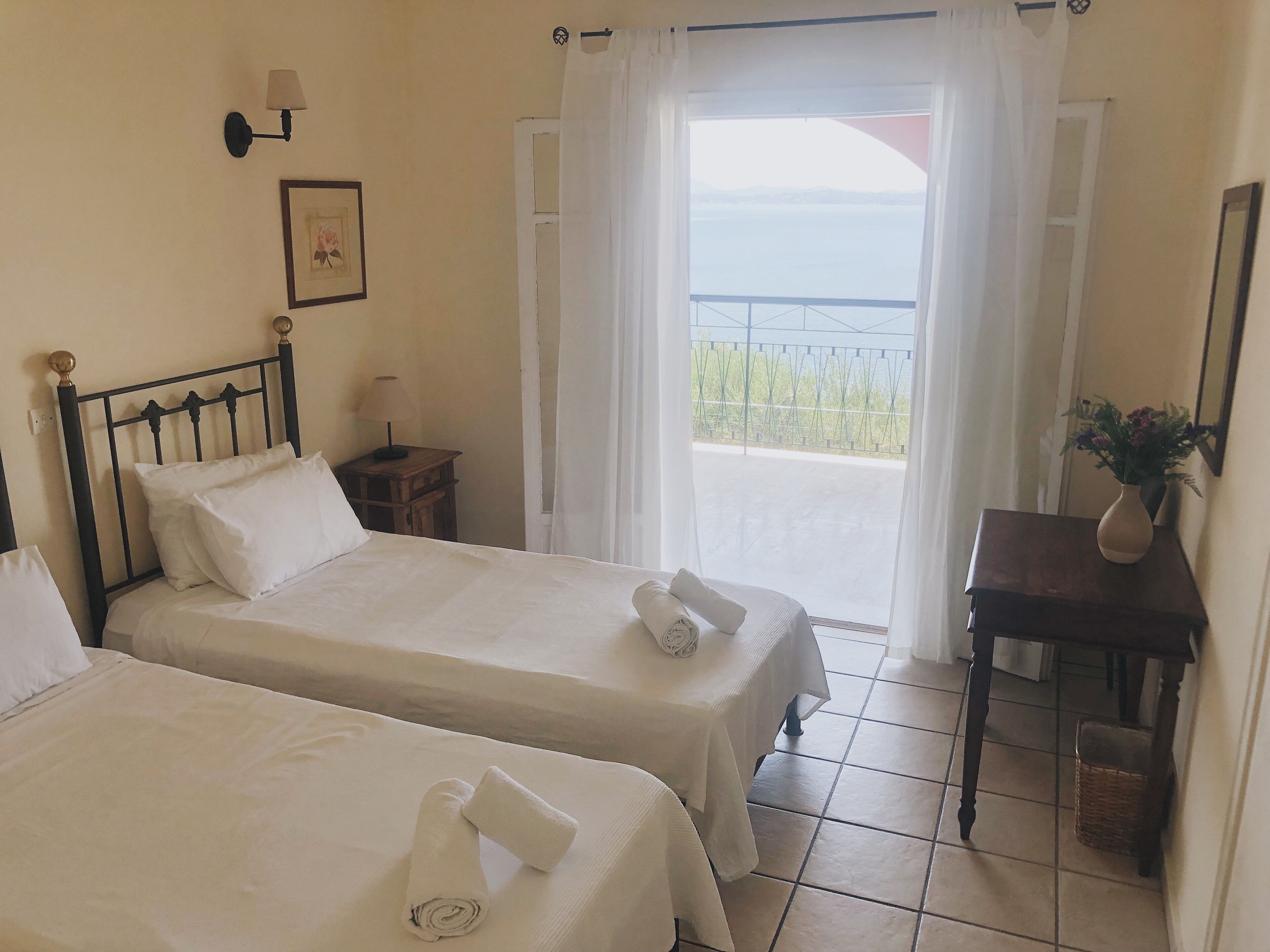 Ein Hotelzimmer mit zwei Betten, einem kleinen Tisch mit einer Vase und Meerblick durch eine Balkontür.
