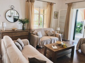 Ein gemütliches Wohnzimmer mit Sofa, Sessel und Couchtisch. An der Wand hängt ein Spiegel, auf einem Tisch steht eine Vase mit Blumen und eine Tür führt auf einen Balkon.