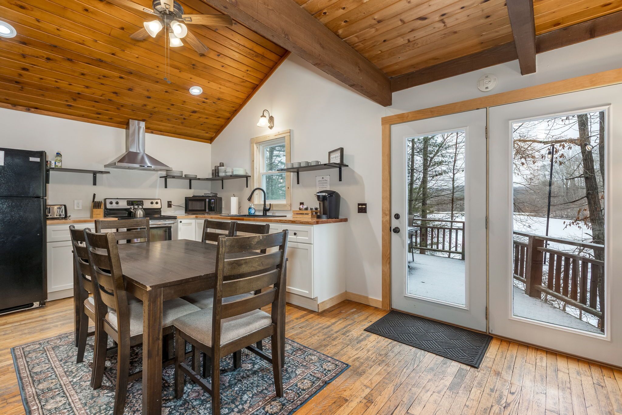 A modern kitchen with wooden ceiling beams, a dining area with a wooden table and chairs, and a view of a snowy deck through glass doors.