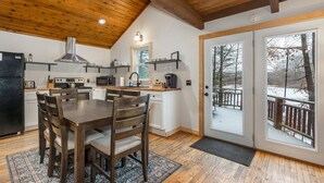 Una cocina moderna con vigas de madera en el techo, un comedor con mesa y sillas de madera, y vistas a una terraza nevada a través de puertas de cristal.