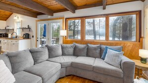 Una sala de estar acogedora con un sofá seccional gris, vigas de madera en el techo y grandes ventanales con vistas al bosque.