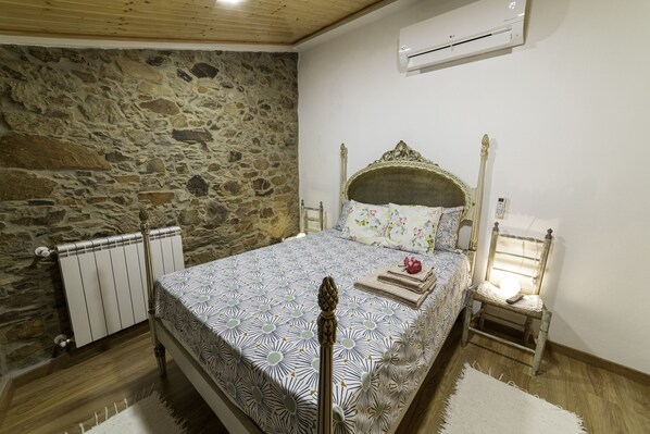 A bedroom with a stone wall, a bed with a patterned bedspread, a radiator, a bedside table with a lamp, and a chair. - Schist House - Panoramic View and Jacuzzi (Sobral Fernando)