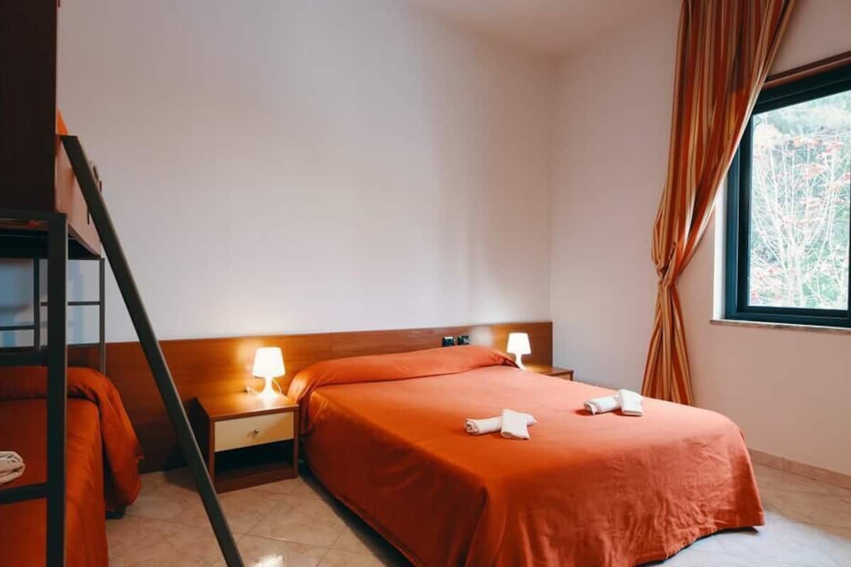 A hotel room with a bunk bed, two bedside tables with lamps, a large window with curtains, and a neatly made bed.