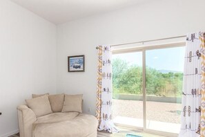 Quarto com sofá bege, uma janela grande com cortinas e um quadro na parede.