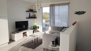 Una sala de estar moderna con un televisor de pantalla plana, un sofá, una mesa de centro y una mesita auxiliar. Hay una ventana con cortinas translúcidas y vistas al exterior.