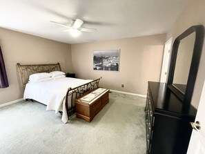 Une chambre à coucher comprenant un lit, une table de chevet, une commode, un ventilateur de plafond et un tableau encadré accroché au mur.