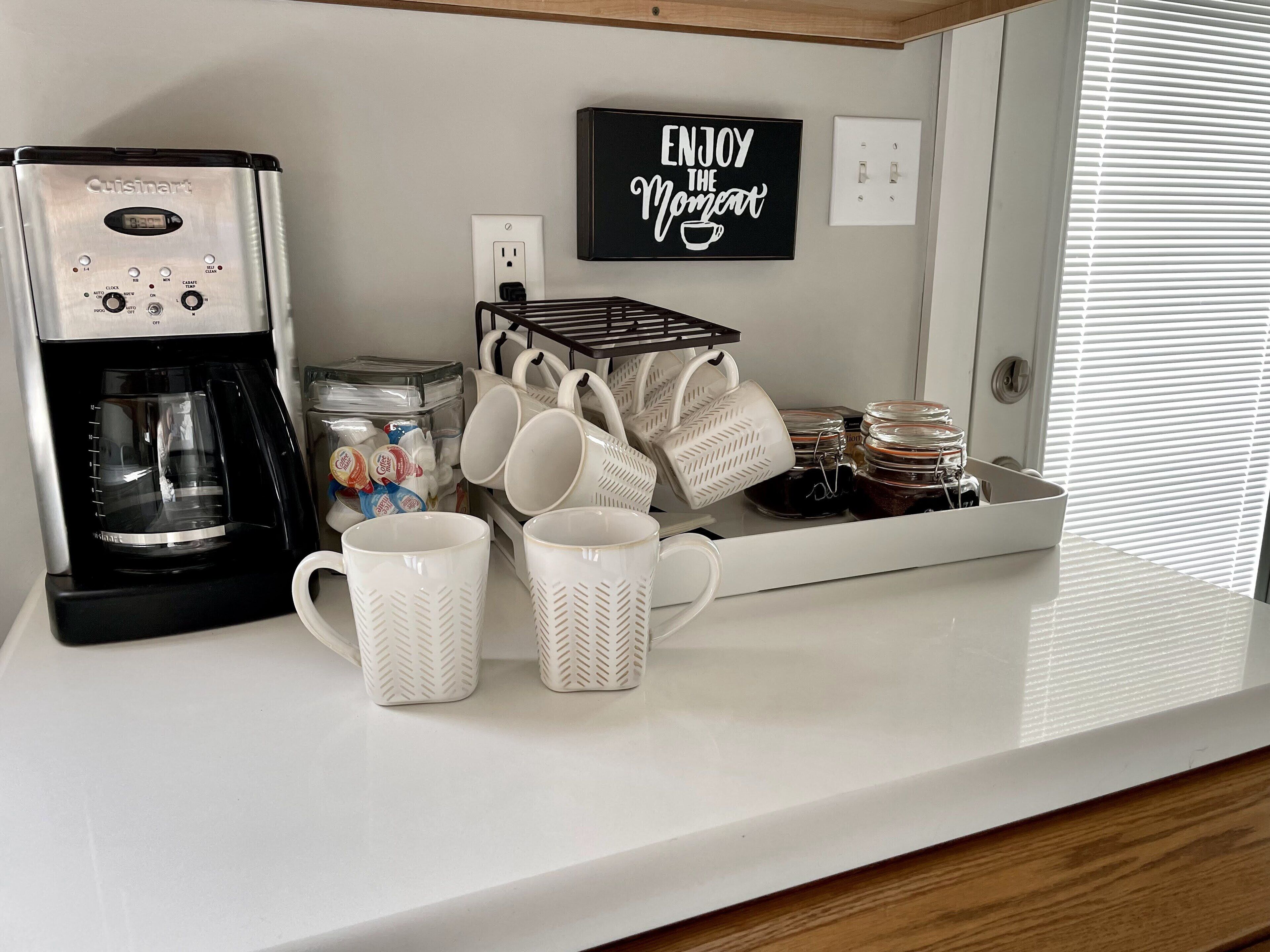 Een kitchenette met een Cuisinart koffiezetapparaat, een koffiehoek, witte mokken, en een bordje met de tekst 'ENJOY THE MOMENT'.