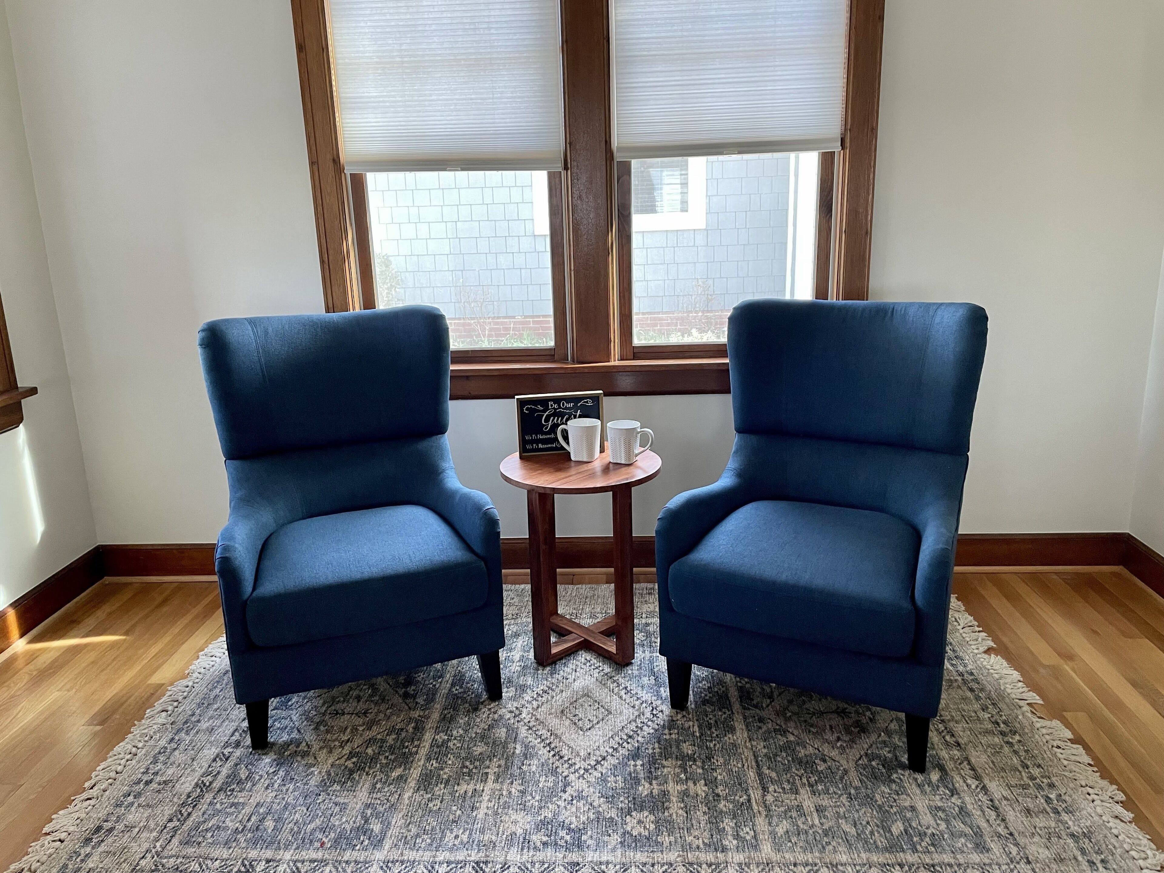 Una habitación con dos sillones azules enfrentados, una mesita de madera con un letrero y dos tazas blancas, y una ventana con persianas.
