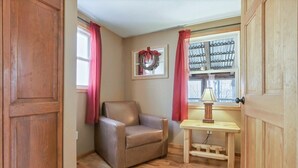 A cozy room with a wooden floor, a brown leather chair, a small wooden table with a lamp, and a window with red curtains. - The Love Shack (Hocking Hills Area) (Rockbridge)