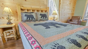 A cozy log cabin room with a bed featuring a bear-themed quilt, a wooden headboard, a leather chair, and a small table with a lamp. - Cliff's Edge (Hocking Hills Area) (Amanda)