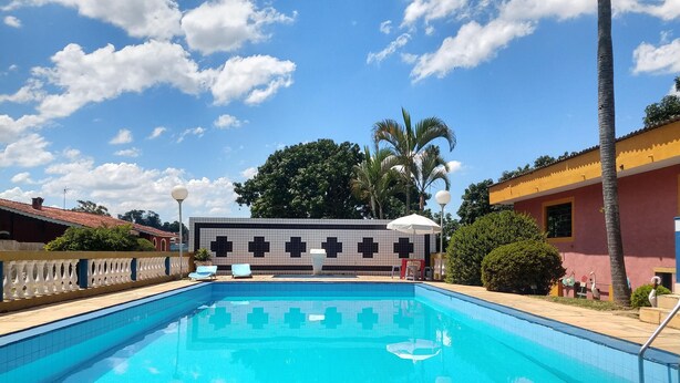 A swimming pool with a tiled deck, lounge chairs, and a building with a cross-patterned wall in the background. - Chácara Sorriso 1 (Atibaia)