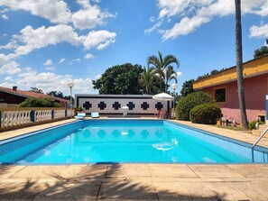 A swimming pool with a tiled deck, lounge chairs, and a building with a cross-patterned wall in the background. - Chácara Sorriso 1 (Atibaia)