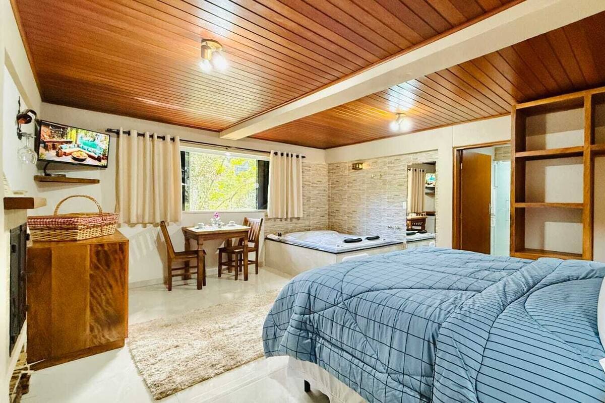Un dormitorio con techo de madera, una cama con colcha azul, una pequeña mesa con sillas, un televisor y una ventana con cortinas.