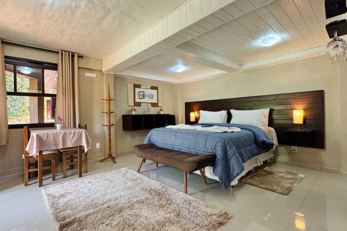 Un dormitorio con cama, mesitas de noche, una banca, una mesa con sillas y un ventanal con cortinas.