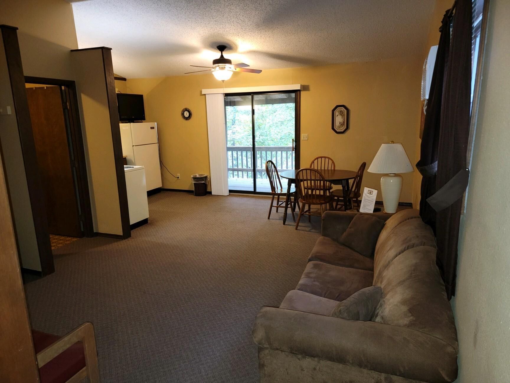 A living room with a sofa, a dining table with chairs, a ceiling fan, and a view through a glass door.