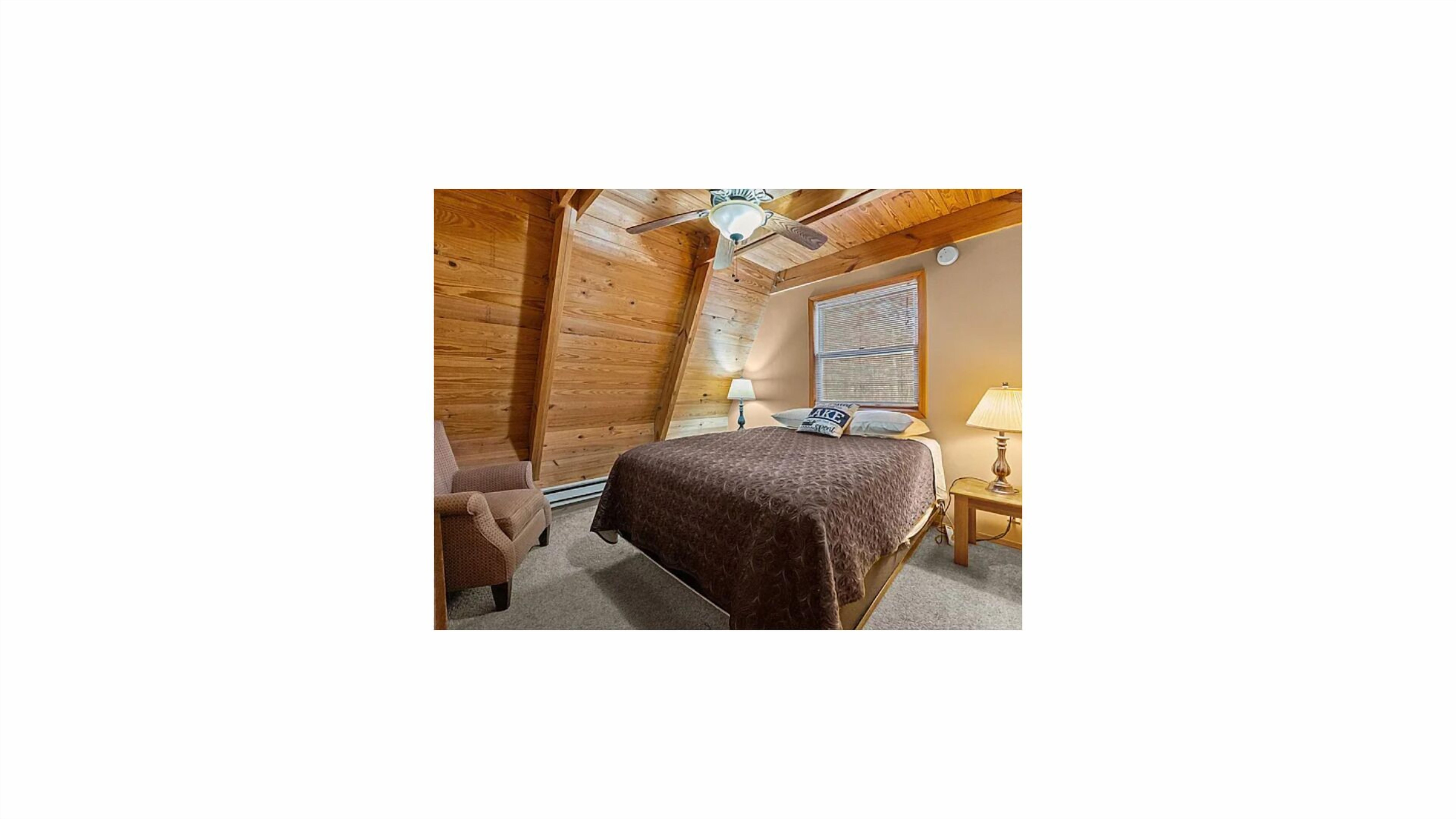 A bedroom with a bed, a chair, a nightstand, a lamp, a ceiling fan, and a window with blinds.