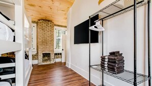 A room with a brick fireplace, wooden flooring, a white wall with a mounted TV, and a metal shelf with folded towels. - Downtown+FREE Parking+Walk Everywhere+Forsyth Park (Savannah)