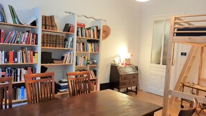 Huoneessa on puinen ruokapöytä, kaksi tuolia, kirjahylly ja pieni työpöytä, jossa on lamppu.
