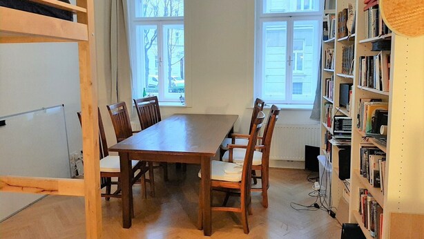 Huoneessa on puinen pöytä ja tuolit, kaksi ikkunaa, joissa on verhot, kirjahylly ja patteri.