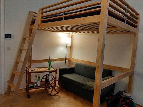 Une chambre avec un lit superposé, une échelle en bois, une petite table avec des bouteilles, un canapé vert et une lampe.