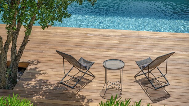 Une terrasse en bois avec deux chaises longues et une petite table ronde, donnant sur une piscine à l’eau d’un bleu limpide.