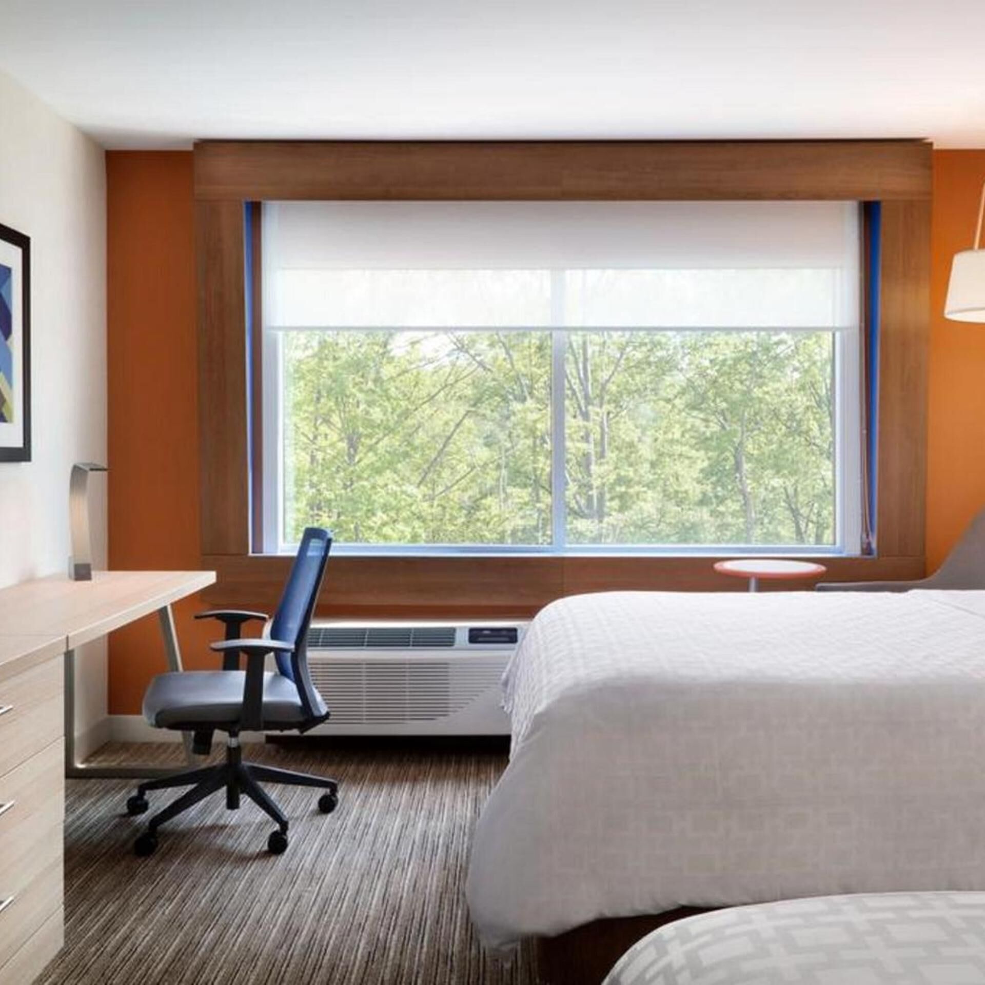 A hotel room with a bed, a desk with a chair, a window with a view of trees, and a wall-mounted lamp.