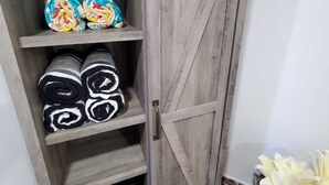 Une armoire rustique en bois, dotée d’une porte coulissante, d’étagères où sont rangés des tapis enroulés, et d’un panier rempli de fleurs blanches.